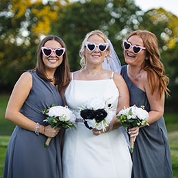 Bridesmaid and bride wearing heart-shaped sunglasses, holding bouquets, smiling in a relaxed outdoor wedding setting.