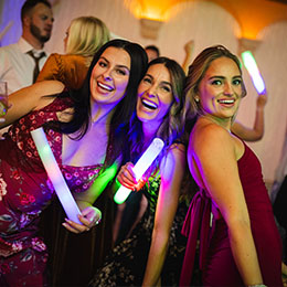 Three women joyfully posing with glowing sticks at a lively event, capturing the vibrant energy and authentic moments of celebration.