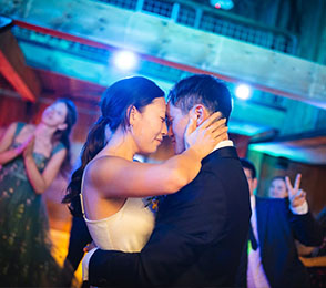 Couple dancing closely at a wedding reception, surrounded by guests celebrating, with vibrant lighting enhancing the candid, joyful atmosphere.
