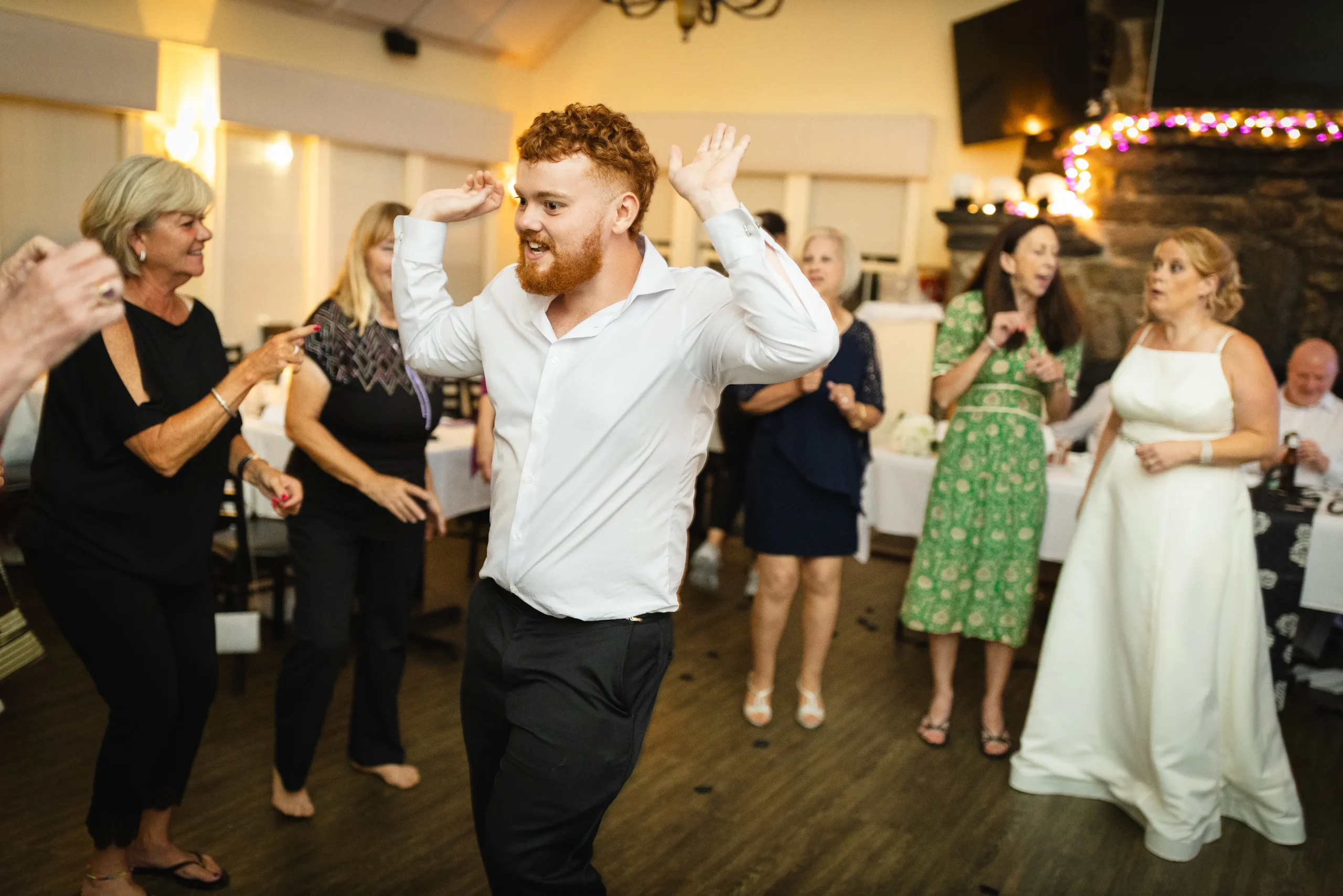 Dancing guests celebrating at a wedding reception, capturing joyful interactions and dynamic movements, with a focus on a man in a white shirt and red hair amidst a lively crowd.
