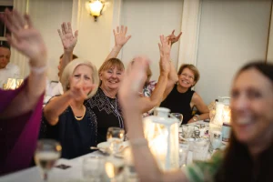 Group of women at a dinner celebration, joyfully raising hands and smiling, capturing candid moments of happiness and connection in a lively event setting.