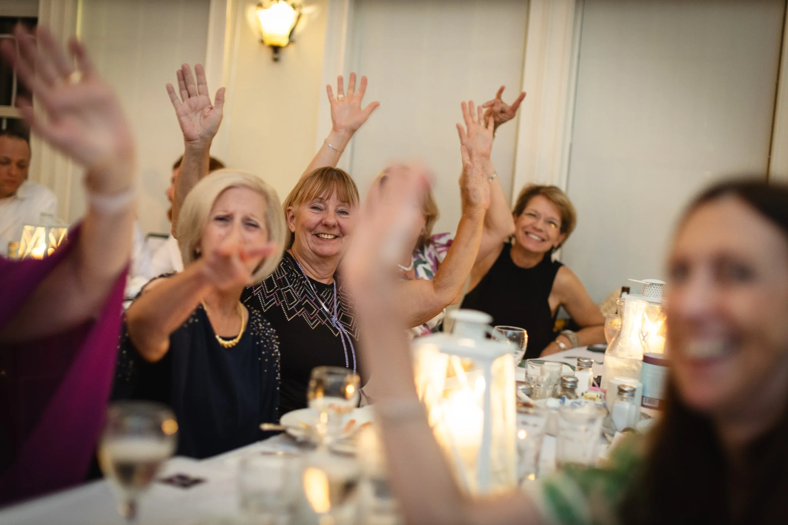 Group of joyful guests raising hands at a wedding reception, celebrating with laughter and excitement, surrounded by a festive dining setup with candles and glasses.