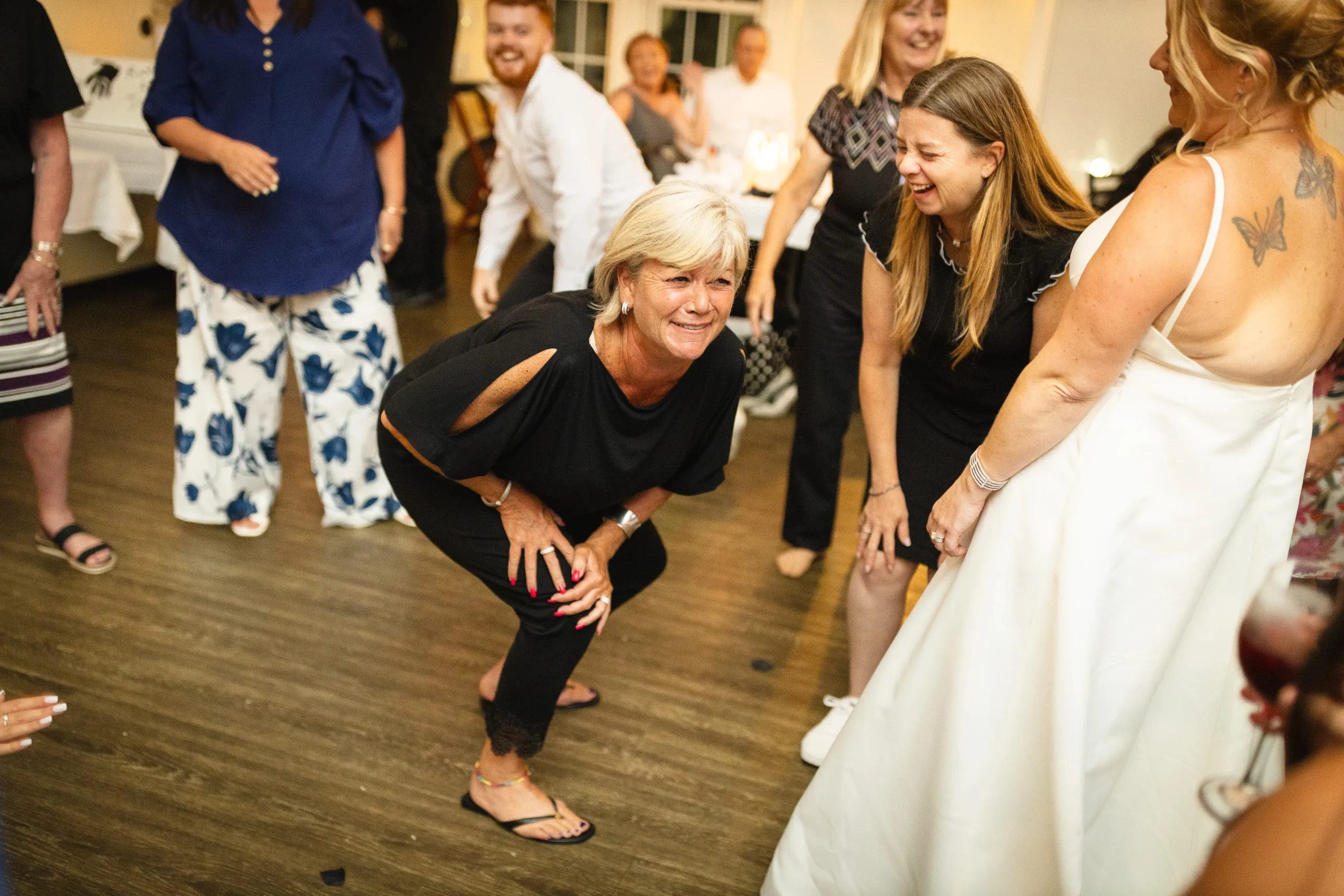Energetic wedding reception scene with guests dancing and laughing, featuring a woman in a black outfit crouching and enjoying the moment, surrounded by others in festive attire, capturing candid interactions and joyful atmosphere.