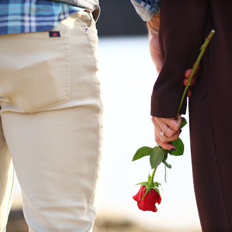 Couple holding hands, one partner presenting a red rose, symbolizing love and connection, in a candid moment by the water, reflecting authentic lifestyle photography.