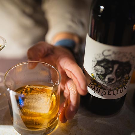 Hand reaching for a glass of whiskey with ice beside a bottle labeled "Dandelion," capturing a relaxed, intimate moment.