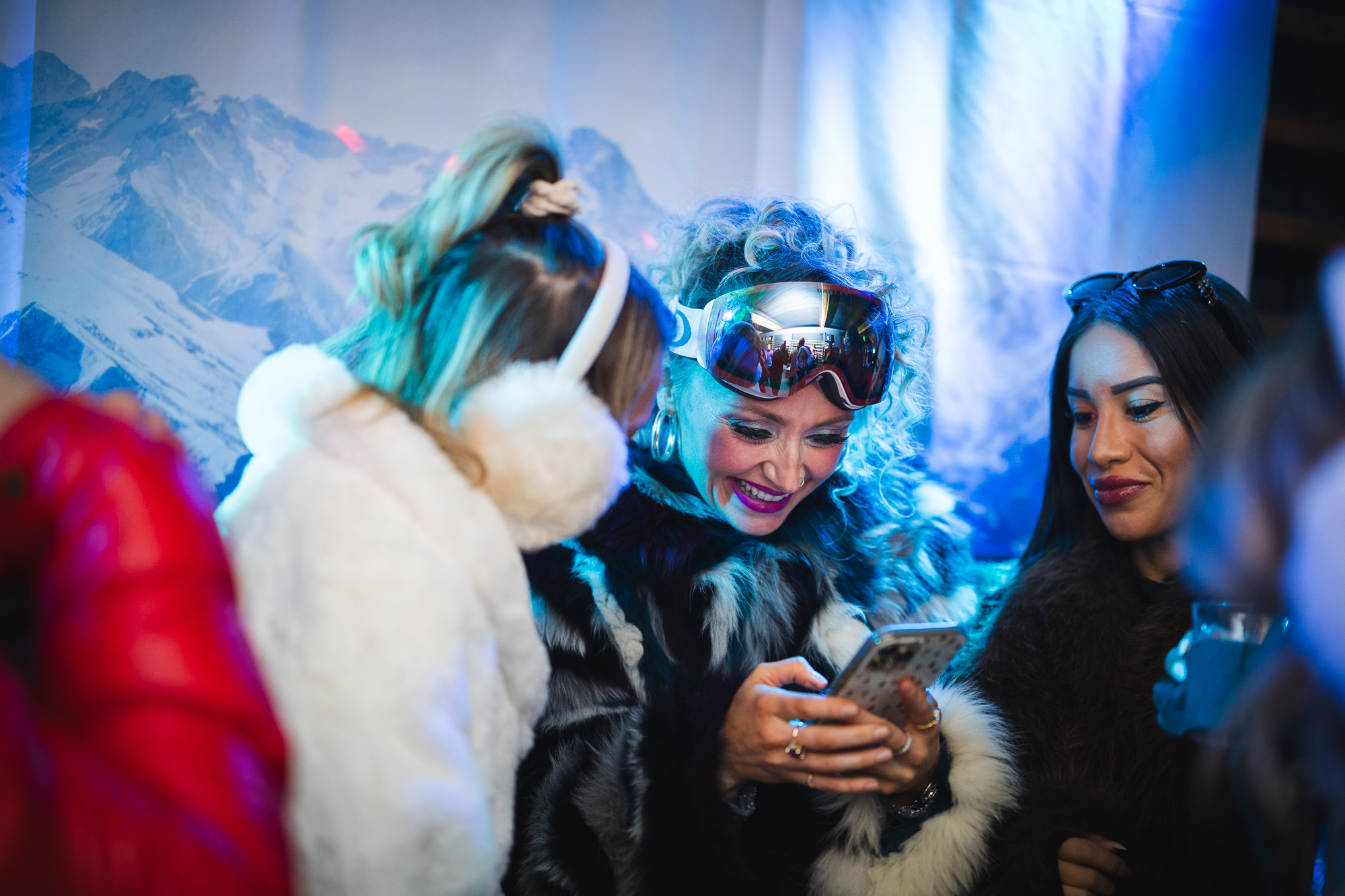 Women in 80's ski chic attire, including retro ski goggles and fur coats, enjoying the Après Ski event at The Bridge At Saugatuck, capturing vibrant social interactions against a snowy mountain backdrop.