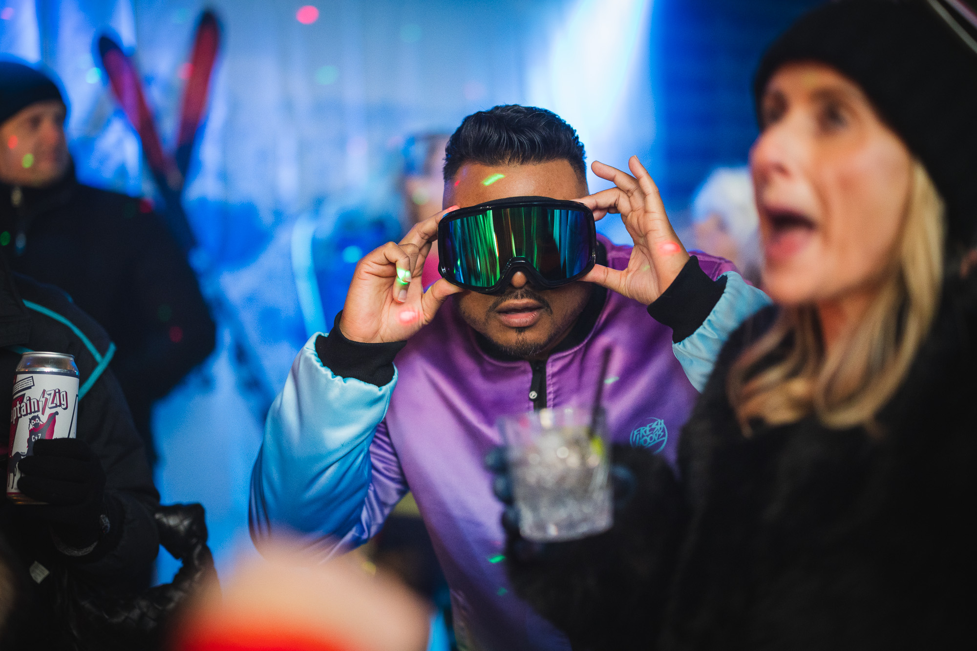 Man wearing retro ski goggles and a colorful vintage jacket at an 80's ski chic themed event, surrounded by festive atmosphere and partygoers.