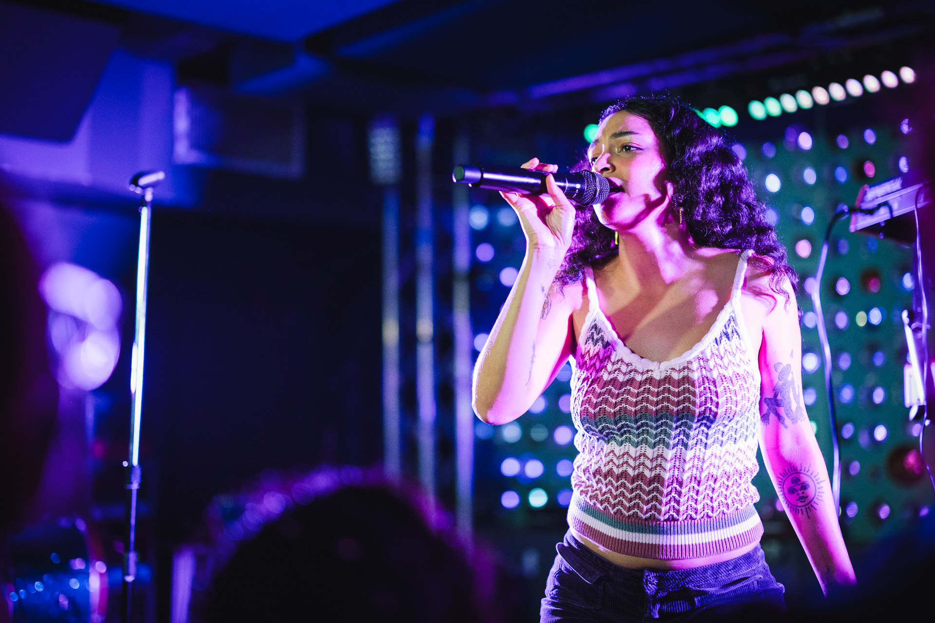 A female artist with dark curly hair sings into a microphone on stage, illuminated by vibrant blue and purple stage lights, wearing a patterned crop top.