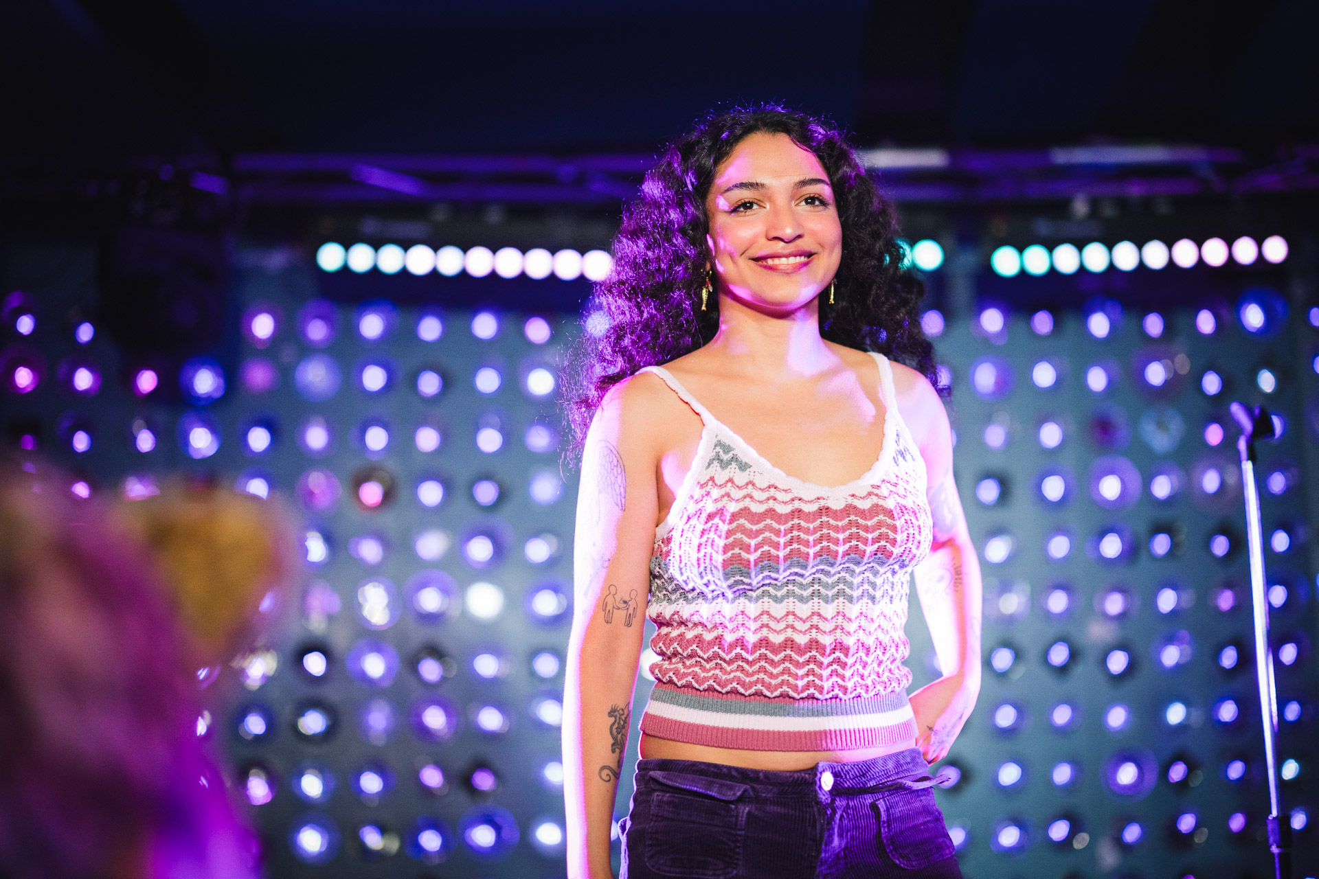 A young woman with long curly hair smiles brightly on a stage, illuminated by purple and blue bokeh lights in the background.