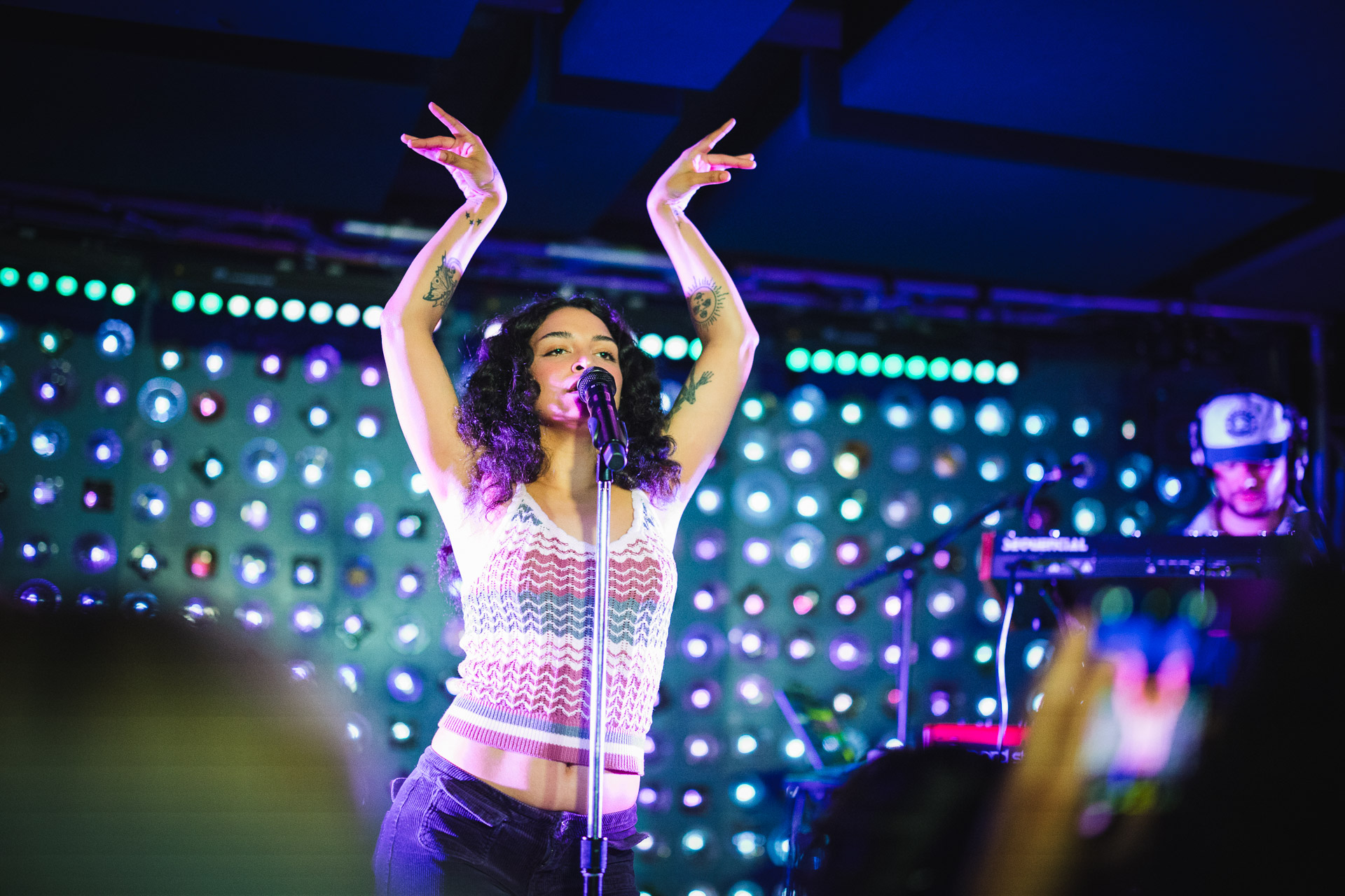 A singer with curly hair and tattoos on her arms performs with a microphone, arms raised, on a stage lit by colorful, bokeh-effect lights.