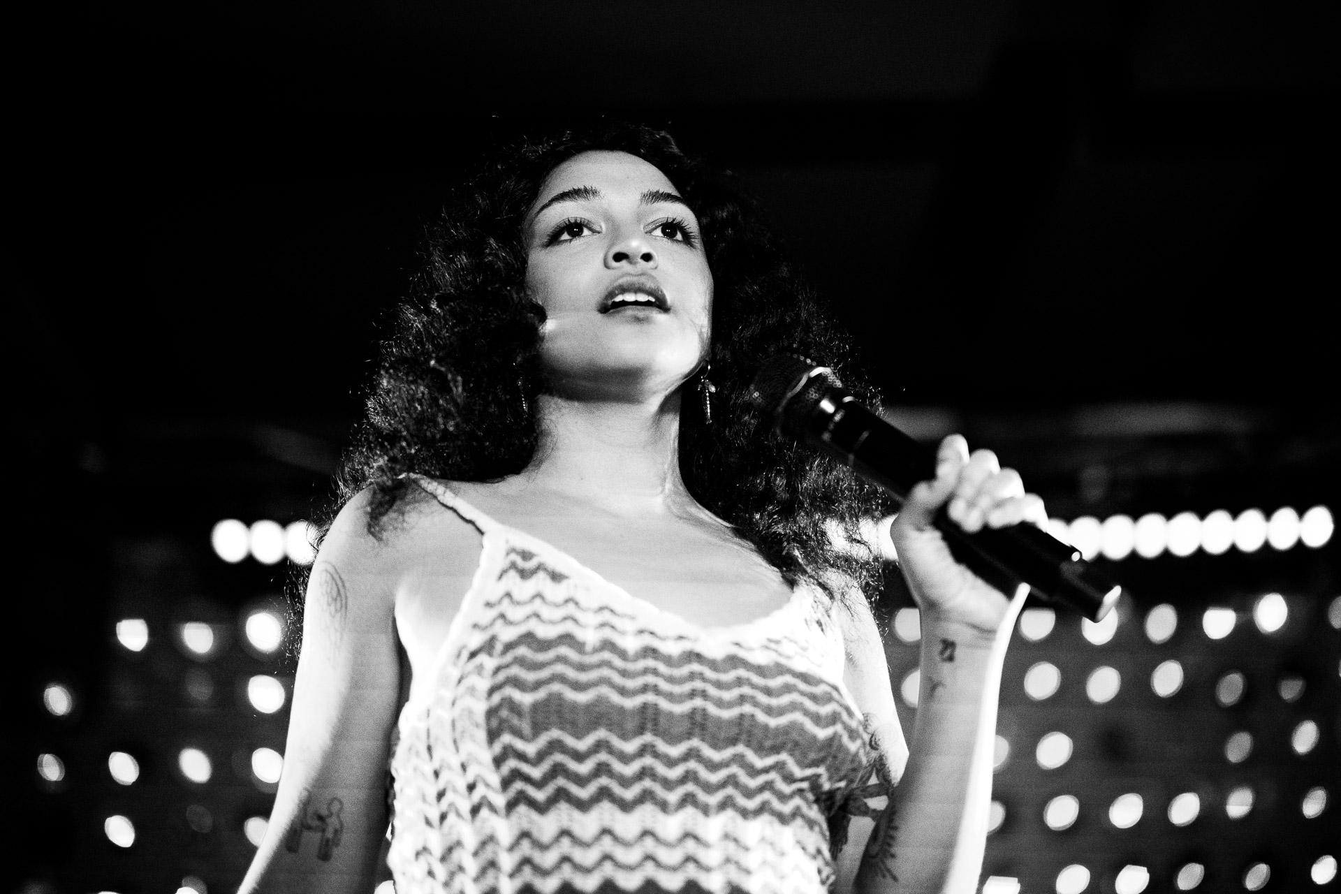 Black and white photo of a female singer with curly hair holding a microphone and looking upwards, performing on stage.