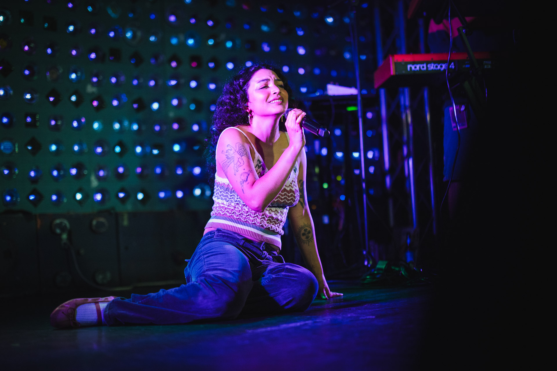 A female performer with curly hair sings passionately into a microphone while kneeling on a dark stage, illuminated by vibrant purple and blue stage lights and a patterned light backdrop.