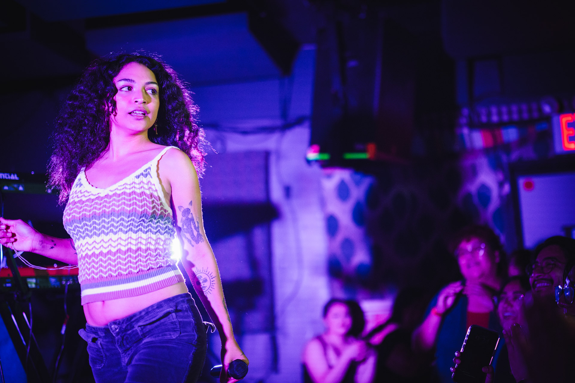 A woman with curly hair, wearing a striped crop top and jeans, holds a microphone on a stage bathed in purple light, with an audience blurred in the background.