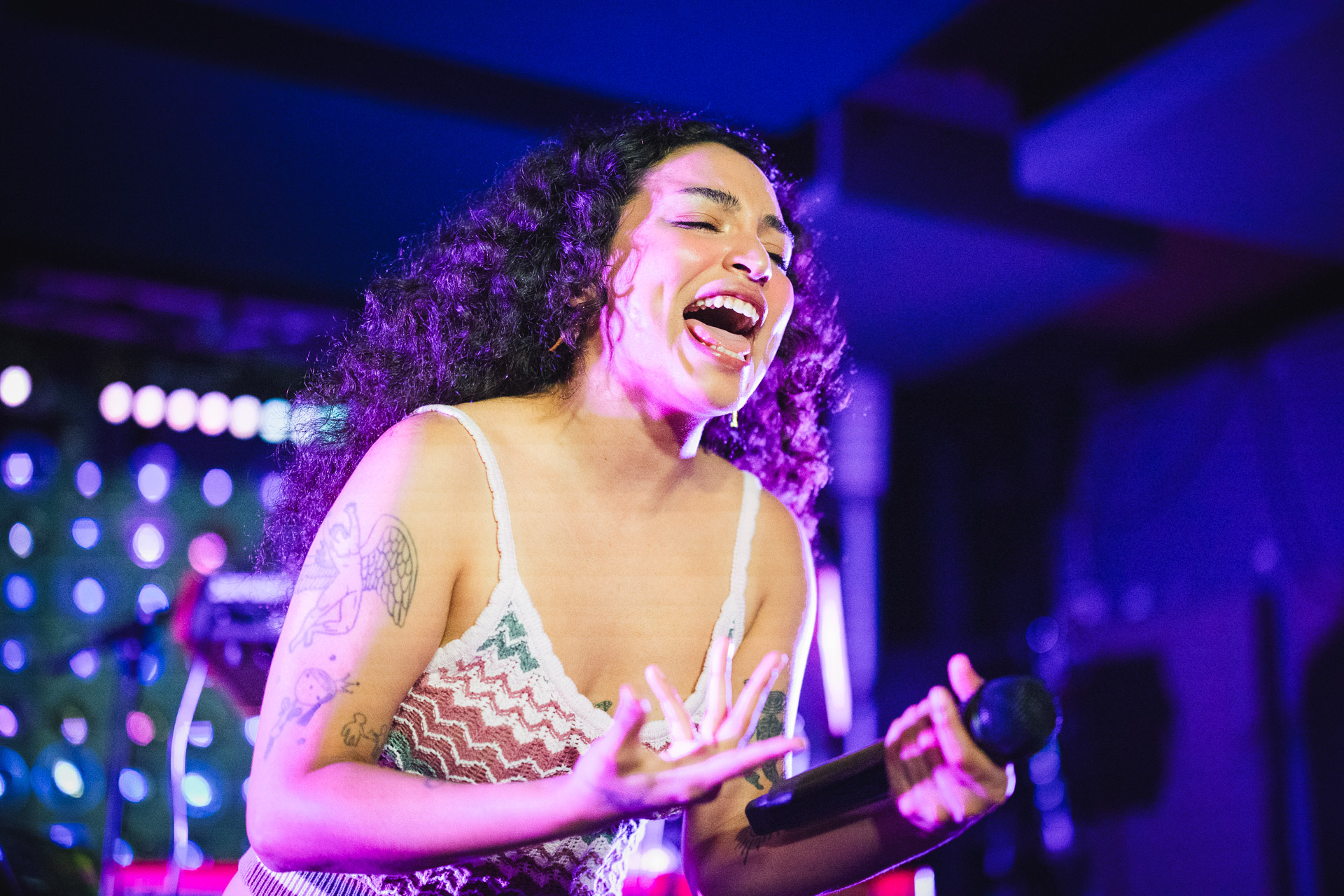 A female singer with dark curly hair and tattoos on her arm, passionately singing into a microphone on stage, illuminated by purple and pink lights.