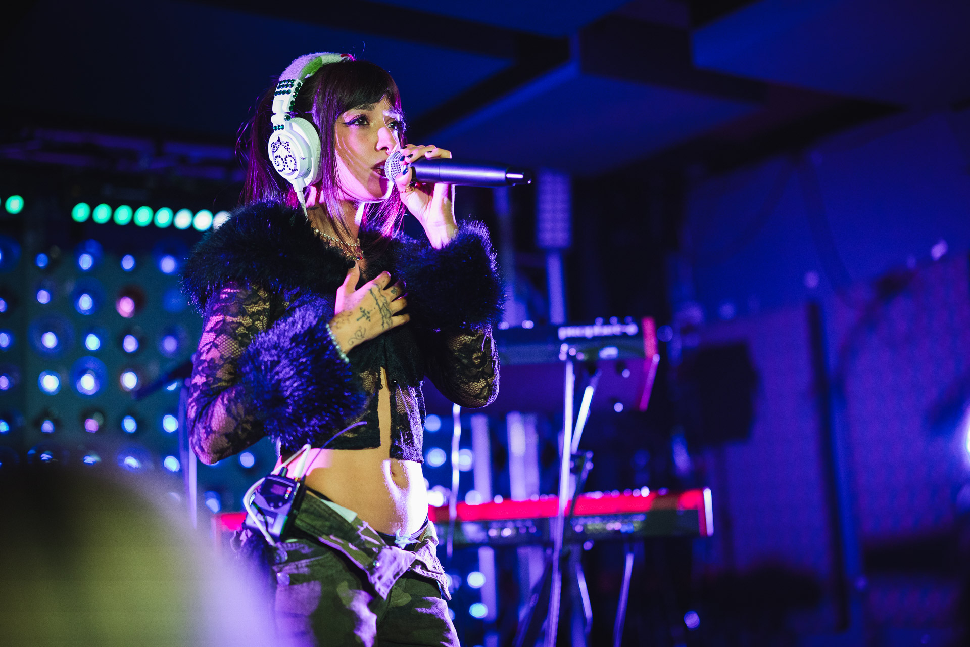 A female musician wearing white headphones and a black lace top sings into a microphone on a vibrantly lit stage.