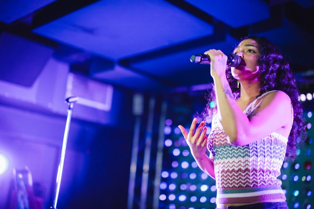 A female singer with long dark curly hair holds a microphone and sings on stage, illuminated by vibrant purple and blue stage lights.