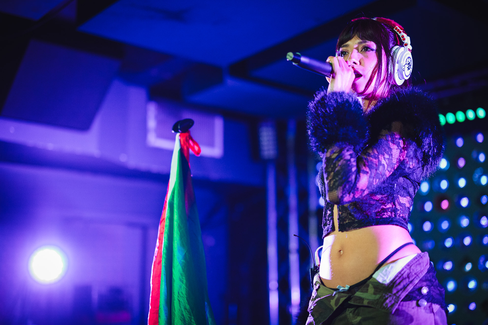 A female vocalist with headphones sings into a microphone on a dark stage lit with blue and purple lights.